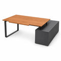 A modern L-shaped desk with a warm brown wood-grain top, charcoal grey, metal frame legs, and a dark gray storage pedestal base, shown on a white background.